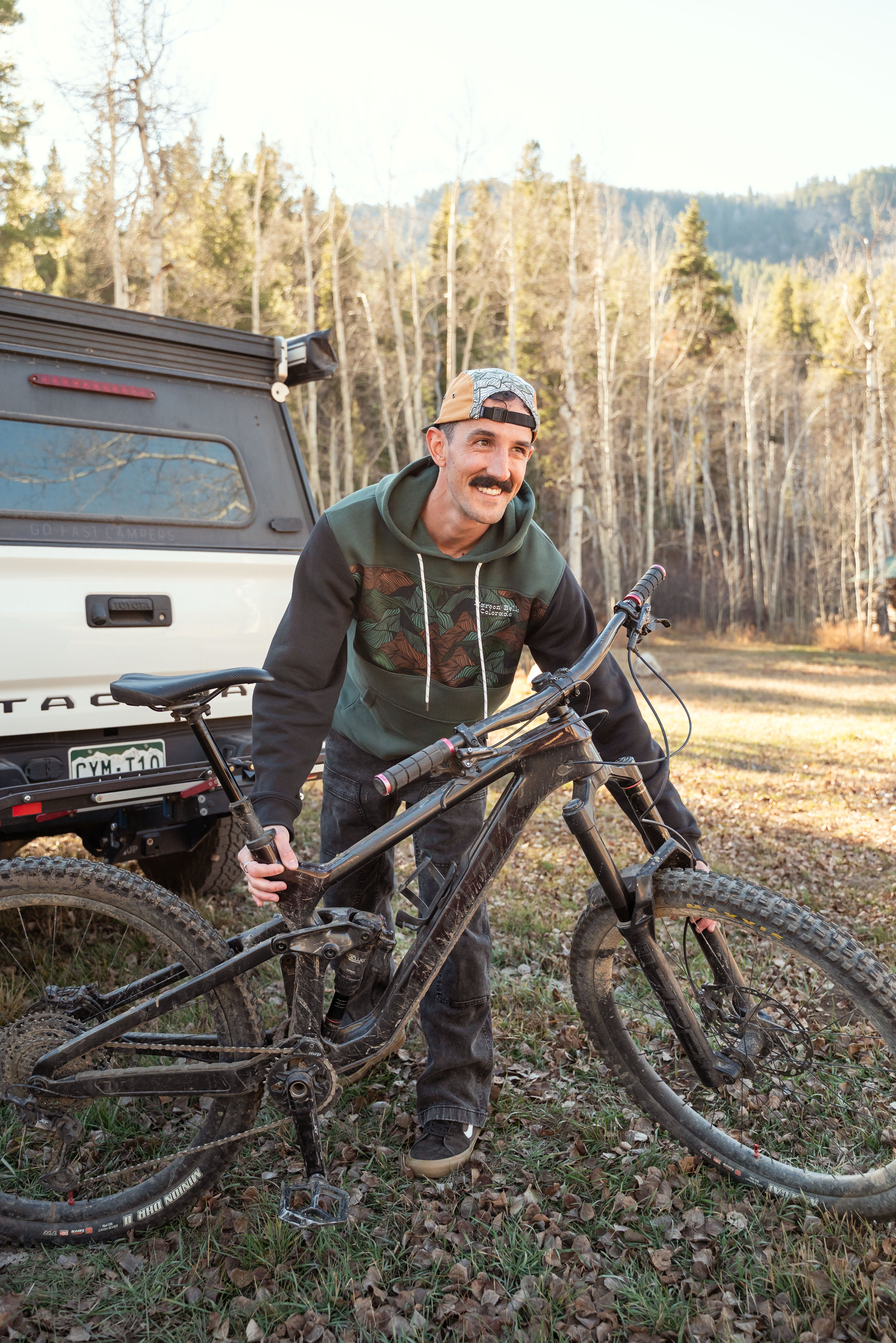Man holding a mountain bike in a forested area with a truck in the background