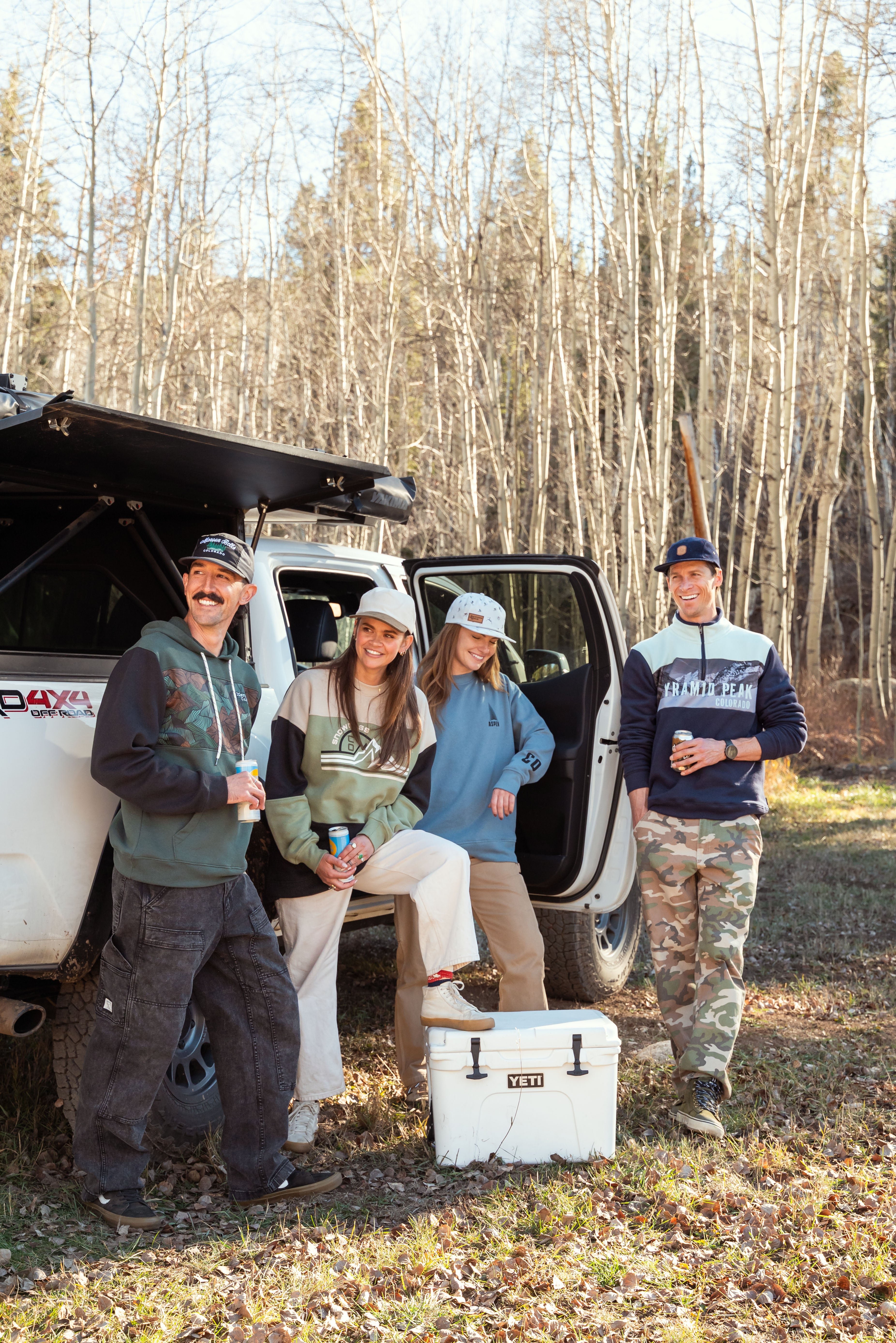 Four people standing near an open car door in a forested area with a cooler and hunting gear.