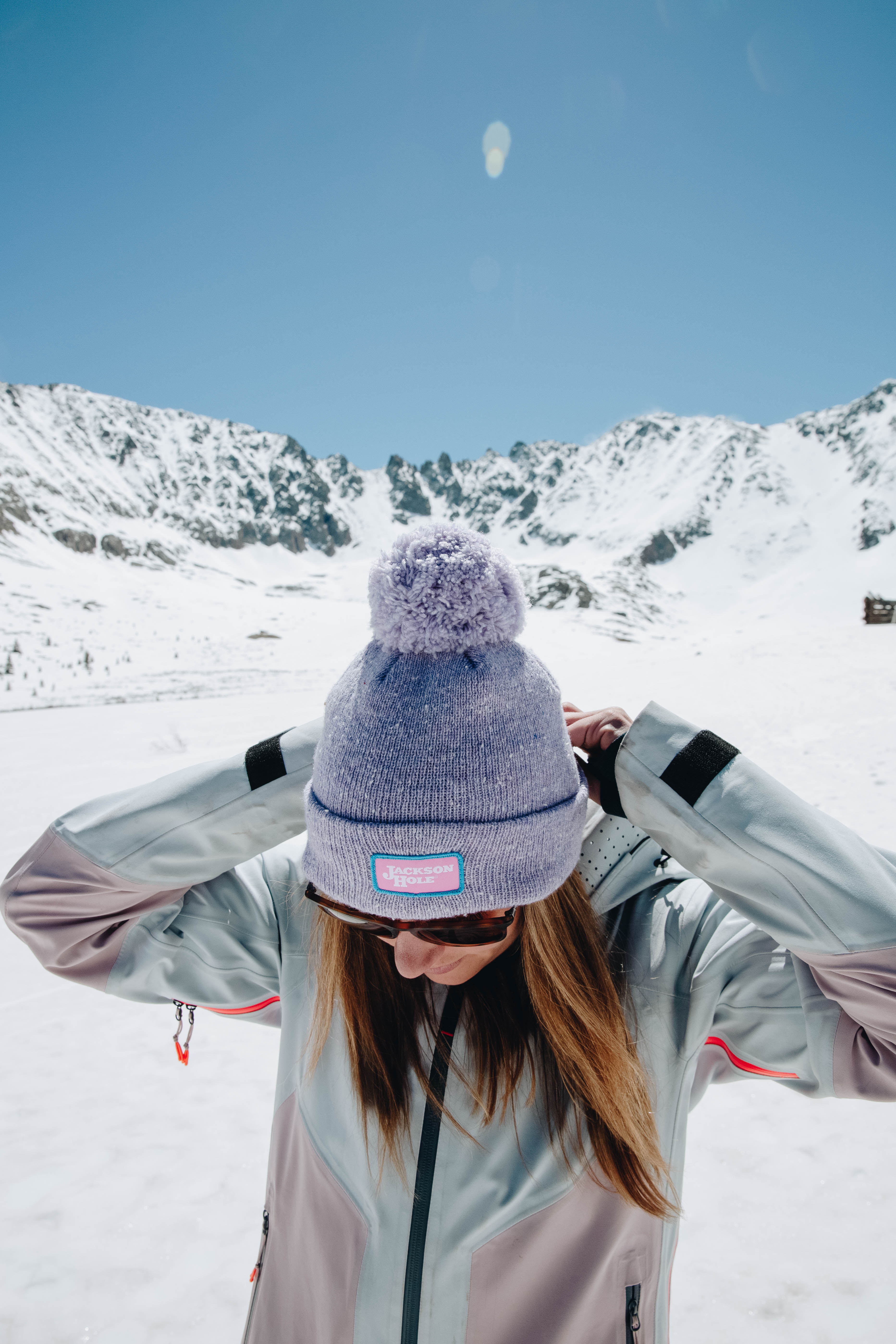 young women on a snowy mountain with both hands on her beanie looking downward and smiling